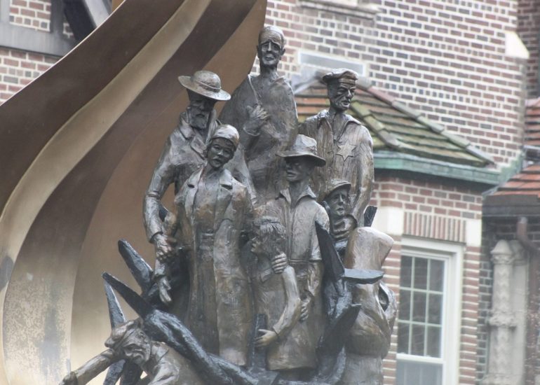 Alfred Tibor : A Life Of Inspiration 6 Bronze statue of multiple figures including men, women, and children in period clothing against brick buildings.