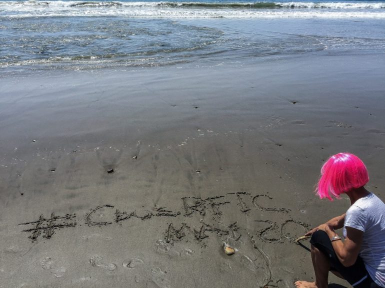 Andrea Cambern Is Coming Home 8 Person with pink hair drawing in sand at a beach with waves in background