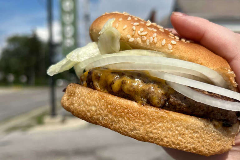 Johnnie's Tavern Is The Best Kept Secret In Columbus 14 Gourmet hamburger with sesame seed bun, grilled patty, mustard, and sliced onions held in hand outdoors.