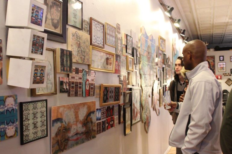 Chomping At The Bit: Debating The Columbus Arts Scene 2 Two visitors view an extensive gallery wall display of framed artwork and prints in an indoor exhibition space.