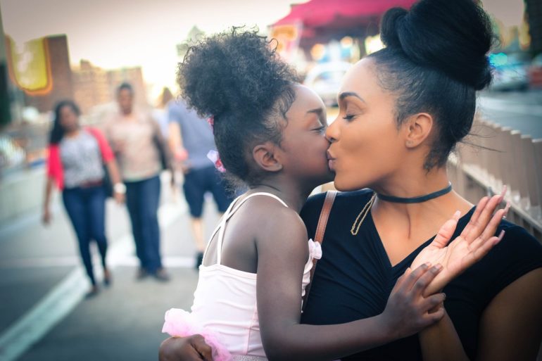 Two women embrace and kiss on an urban street with pedestrians in the background.