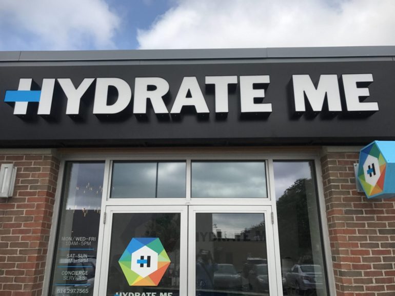 First I Got Super Drunk, Then I Stumbled Into A Hydration Clinic The Next Day. Here’s What Happened. 11 Storefront with "Hydrate Me" signage, brick facade, white doors, and colorful hexagonal logo.