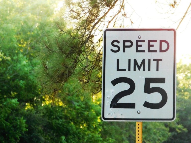 Here Are The Worst Speed Traps In Franklin County 1 A 25 mph speed limit sign mounted on a yellow post surrounded by green vegetation and tree branches.