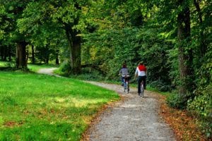 Two cyclists ride on a tree-lined gravel path through a green forest park.