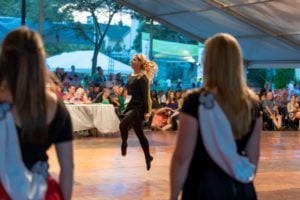 Dancer in black outfit performs on stage as audience watches from tent venue at dusk.
