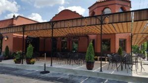 Outdoor patio with red brick building, wooden shade structure, and dining furniture.