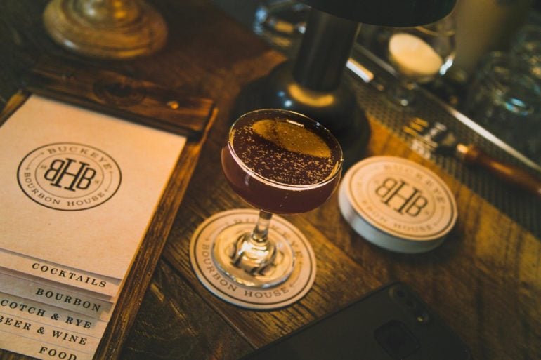 The Best Cocktail Bars In Columbus 3 Dark cocktail with cocoa powder garnish on wooden bar with branded coasters and menu.