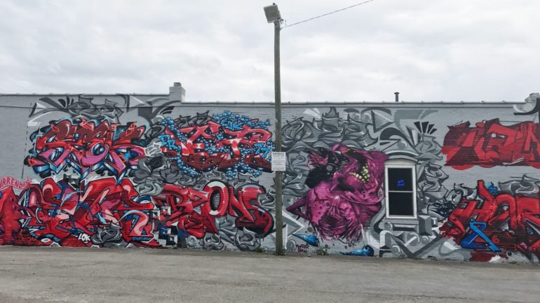 A Controversial Clintonville Mural Is Being Repainted... Again 2 Vibrant red and blue graffiti art covers an industrial building wall under cloudy sky.