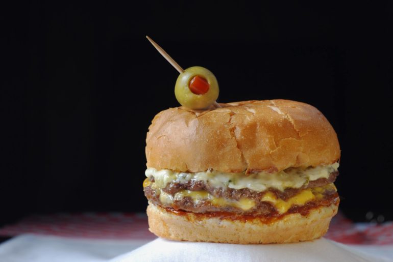 Why Are People Going Crazy Over Swensons Drive-In? 1 Gourmet cheeseburger topped with olive and pimiento garnish on black background.