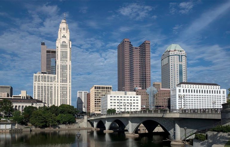 The 10 Tallest Buildings In Columbus, Ohio (And What’s Next) 3 Downtown skyline featuring historic tower, modern buildings, and arched bridge over river.