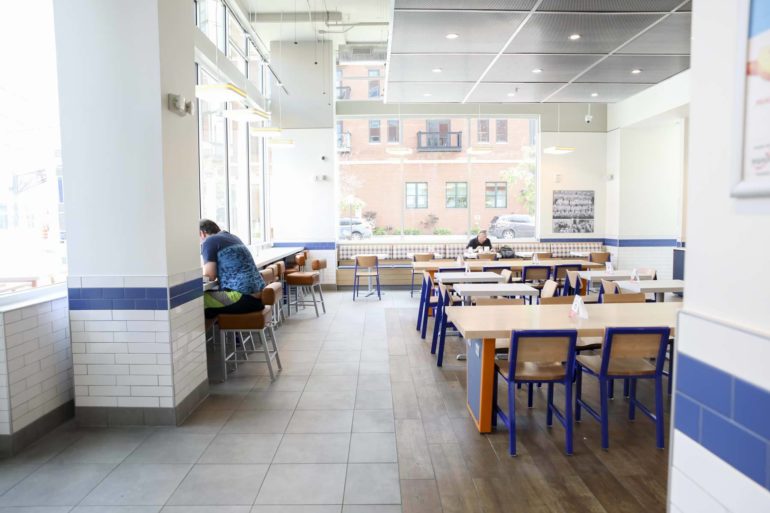 Modern cafeteria interior with blue chairs, wooden tables, and large windows overlooking brick buildings.
