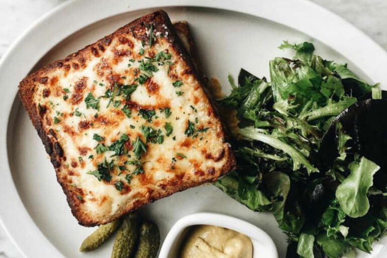 Gourmet cheese toast with herbs, green salad, and pickles on white plate.