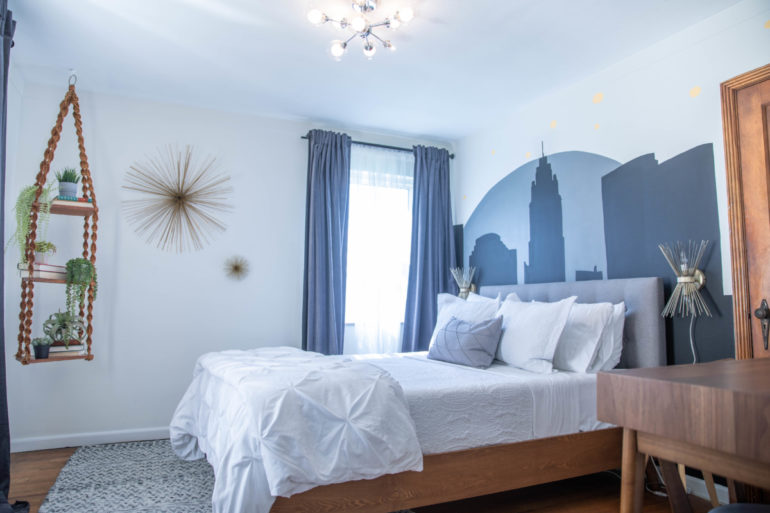 Bedroom with white bedding, blue curtains, a Columbus skyline mural, and hanging plant shelf