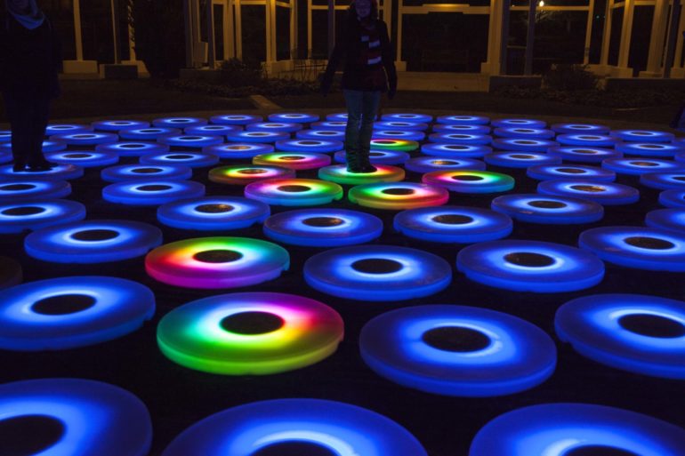 The Interactive Light Installation At Conservatory Aglow Is Pure Joy 18 Illuminated blue and rainbow-colored circular lights on dark ground with people standing nearby at night.