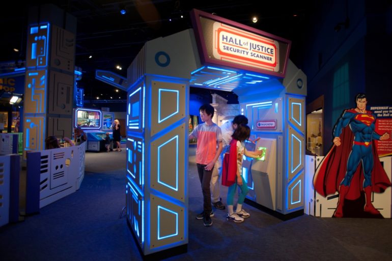 Holy Jackpot, Batman! An Awesome DC Super Heroes Exhibit Is Coming To COSI 2 Interactive "Hall of Justice Security Scanner" exhibit with neon blue lighting and visitors inside futuristic-styled booth.