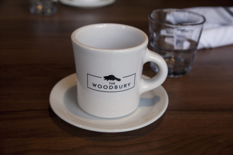 White coffee mug with "The Woodbury" logo and saucer on dark wooden table.
