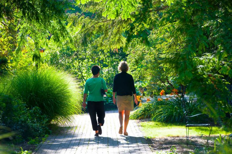 Two women walking along a shaded garden pathway with lush greenery