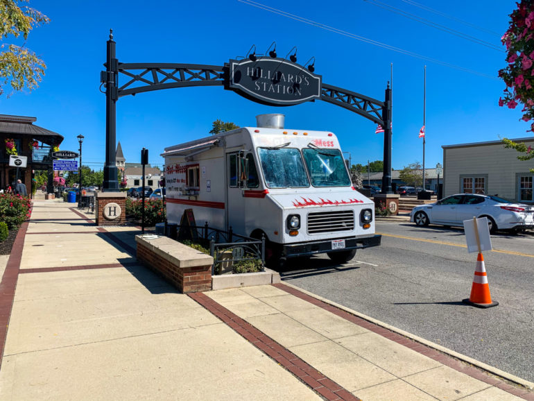 Hot Mess Food Truck Is Anything But A Hot Mess 5 Food truck parked under ornate black sign reading "Hilliards Station" on sunny street with brick buildings.
