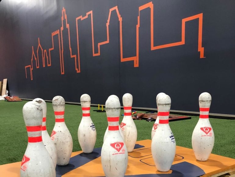 A 50,000+ Square Foot Indoor Adventure Park Is Open In Worthington 7 Bowling pins with red stripes arranged on orange mat against dark wall with neon skyline outline.