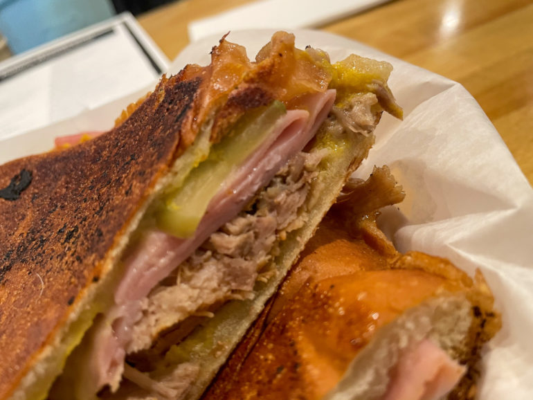 Close-up of a toasted sandwich filled with meat, ham, and pickles on white plate.