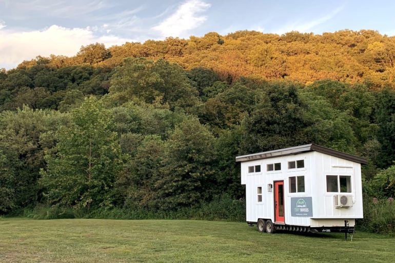 The Gladiola tiny house exterior on a trailer in a grassy clearing near autumn trees