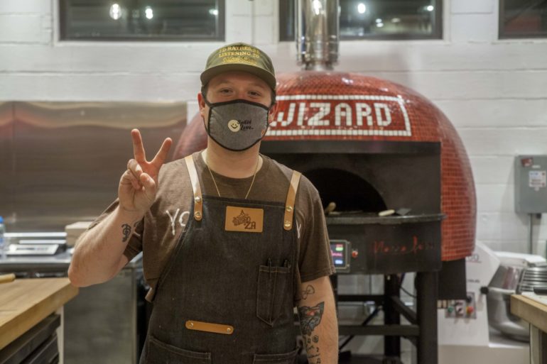 We're Off To See The Wizard Of Za 1 Man making peace sign in front of red pizza oven in commercial kitchen.