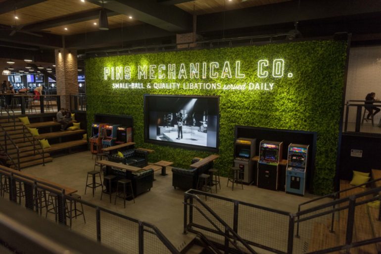 PINS + 16-Bit At Easton Is Open And It's Absolutely Massive 10 Vintage arcade games and seating area beneath neon-lit green wall signage in modern entertainment venue.