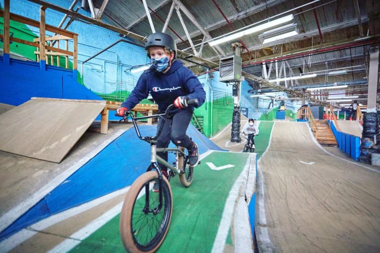 The World’s Longest Indoor Mountain Bike Trail Is Right Here In Ohio 8 Person riding BMX bike indoors on blue and green track wearing helmet and mask.