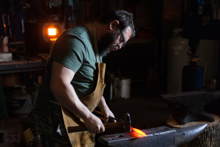 Try Your Hand As A Blacksmith With Columbus Forged In Fire Contestant 1 Blacksmith wearing protective gear works with glowing hot metal at anvil in workshop.