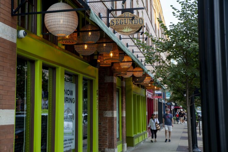 Vibrant lime green storefront with woven pendant lights and "Now Hiring" sign on urban street.