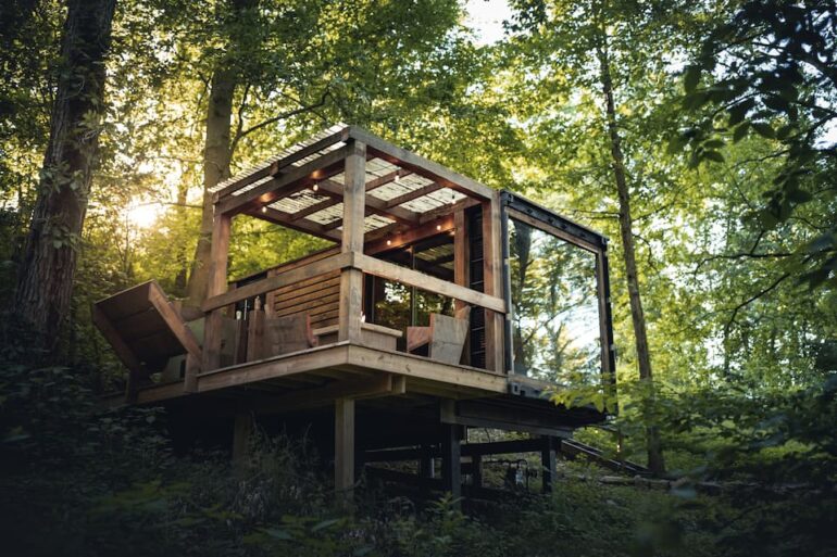 This Storage Container Tiny Home Is Bringing Modern Design To Amish Country 12 Modern elevated wooden cabin nestled among trees in a forest setting.