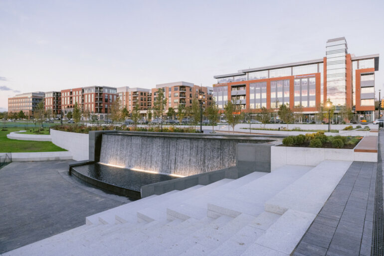 The Gorgeous New Riverside Crossing Park Is Open In Dublin 6 Modern waterfront plaza with water feature, landscaping, and mixed-use residential buildings at dusk.