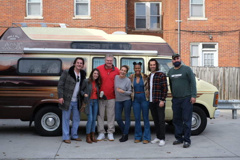 Seven people pose together in front of a vintage two-tone bus on an urban street.
