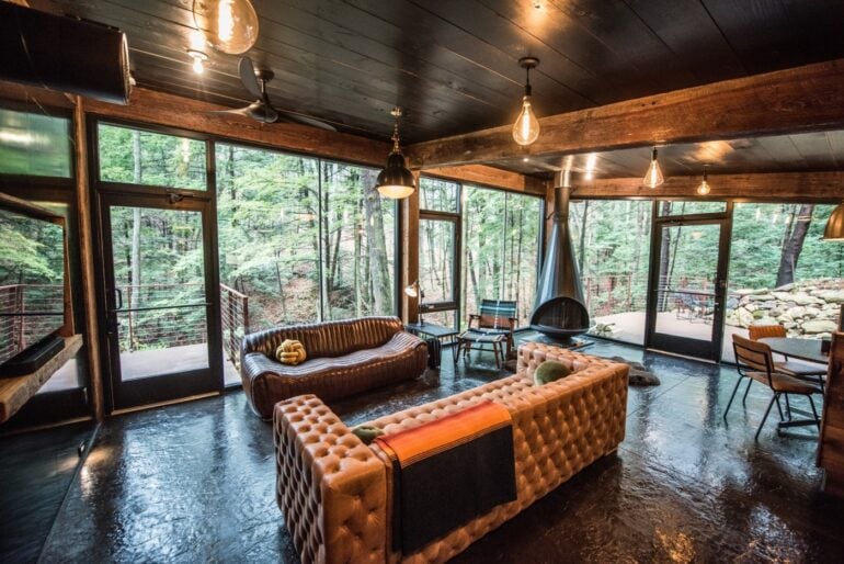 This Frank Lloyd Wright Inspired Cabin Lets You Sleep Above A 50-Foot Cavern 2 Luxury modern cabin interior with leather seating, fireplace, and forest views through expansive windows.