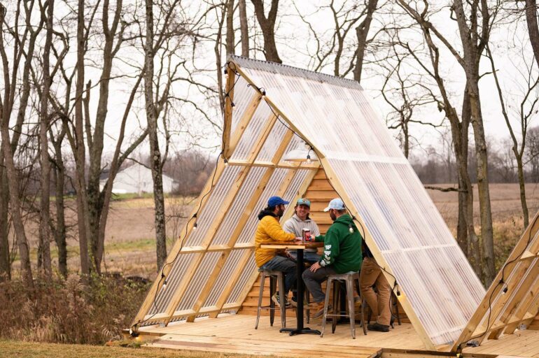 Three men sit at a table inside a modern A-frame structure in a wooded area.