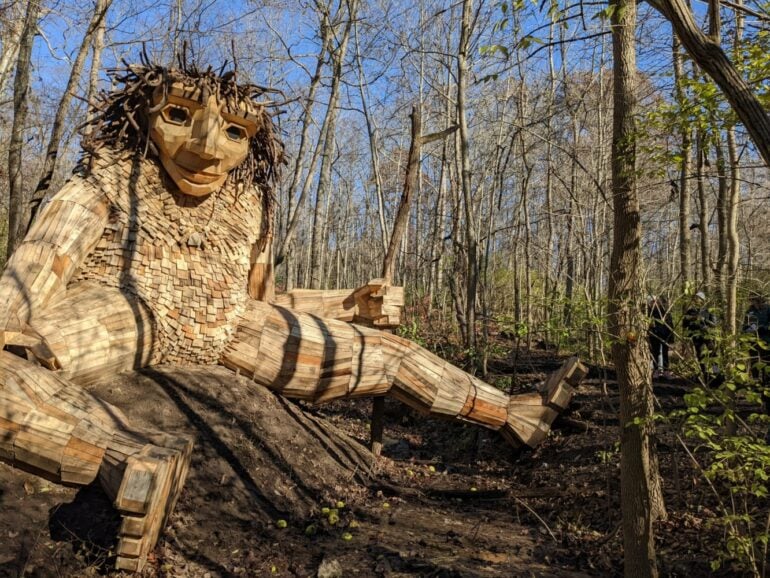 Giant wooden troll sculpture reaching across a wooded trail