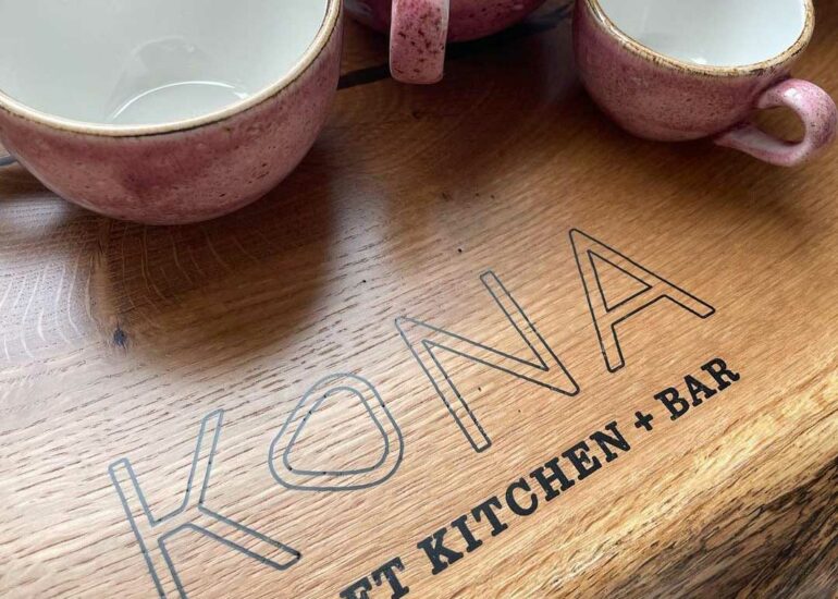 A New Kona Coffee-Inspired Eatery Will Open At Bridge Park Next Month 4 Pink ceramic mugs sit above engraved wooden cutting board reading "KIONA KITCHEN + BAR"