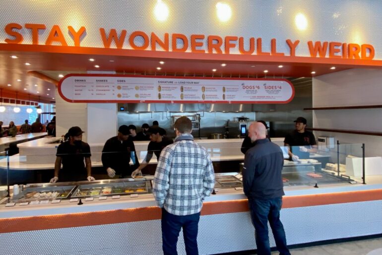 Weenie Wonder Opens Today At Bridge Park 2 Interior of casual fast food restaurant with orange signage and customers at counter ordering food.