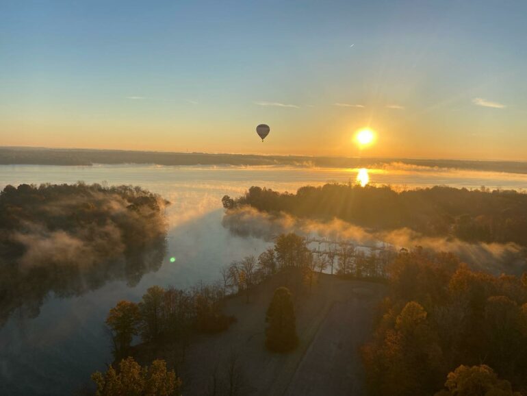 Hot air balloon floats above misty river valley at sunrise over forested landscape.