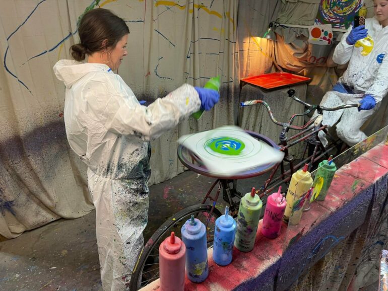Woman in protective gear uses spinning bicycle wheel for colorful paint art creation.
