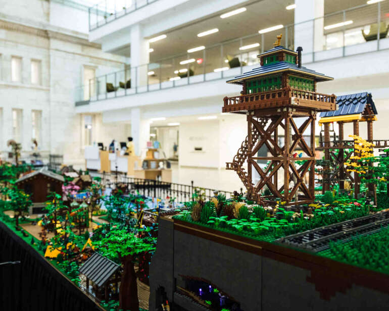 Detailed close-up of elaborate LEGO watchtower structure with blue roof in large indoor display, modern building atrium visible behind.