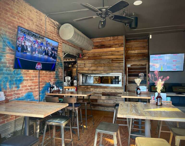 Modern restaurant interior with wooden tables, bar counter, exposed brick walls, and menu displays.