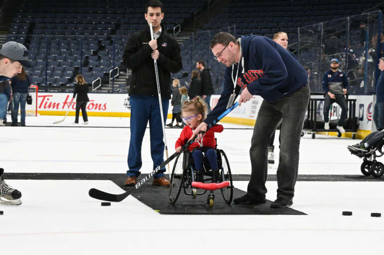 A Kid Again Is Changing Lives For Families Of Kids With Life-Threatening Conditions 7 Young child in wheelchair plays adaptive ice hockey with adult instructor on arena ice.