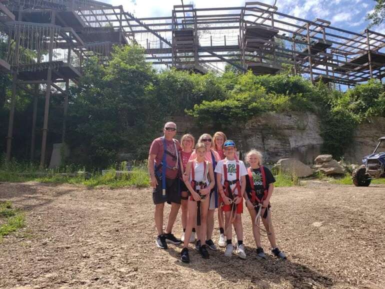 The World's Largest Family Ropes Course Is Tucked Away In Hocking Hills 3 Family group posing on dirt ground with industrial metal structures and overgrown vegetation visible behind them.