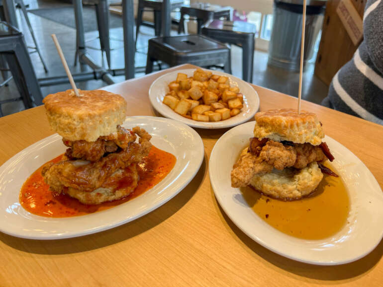 Two biscuit sandwiches with meat and sauce, served with crispy potatoes on wooden table.