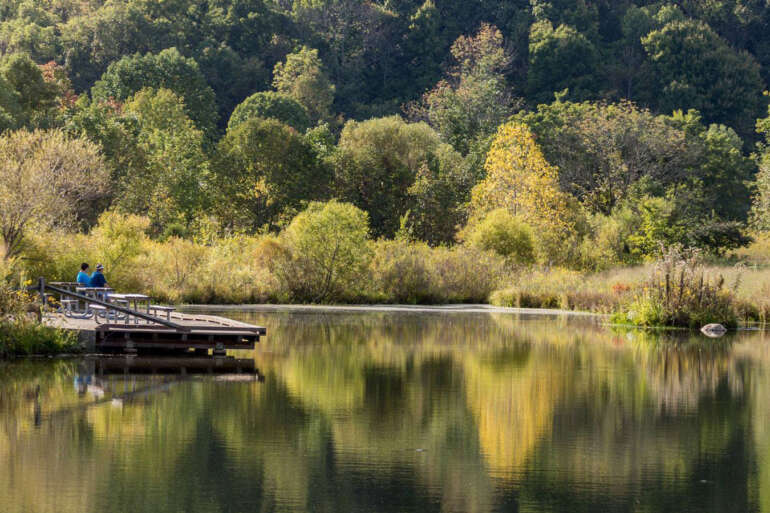 Two anglers stand on a dock over a pond reflecting autumn trees