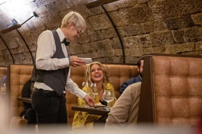 Server presenting food to smiling woman at upscale restaurant booth with stone walls.