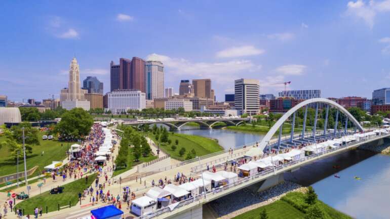Here’s How To Get VIP Access At The Columbus Arts Festival This Summer 10 Wide aerial view of riverfront festival with crowds, modern arch bridge, and downtown skyline under blue sky.