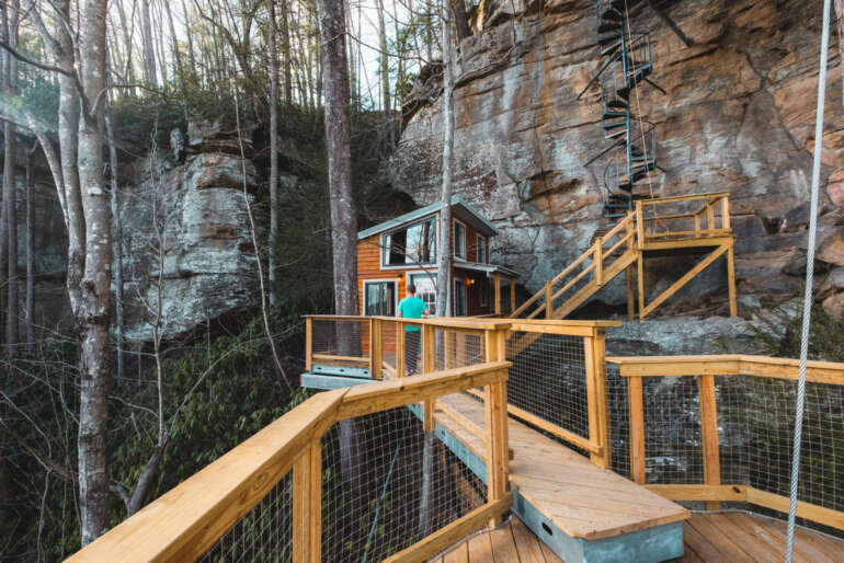 This Stunning Treehouse Perched On A Sandstone Cliff Will Take Your Breath Away 2 Wooden deck with metal railings overlooking treehouse cabin built into rocky cliff face.