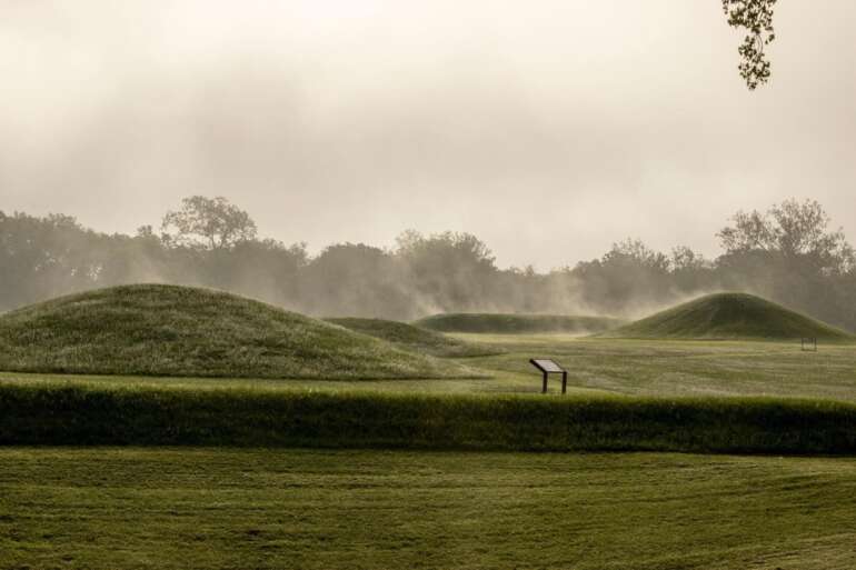 Misty morning view of ancient earthen mounds with interpretive sign in grassy field.